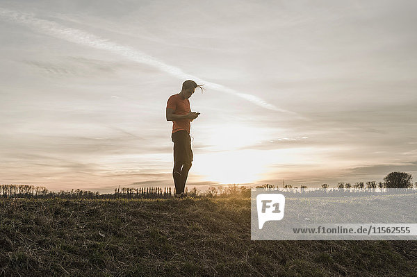 Sportler beim Blick auf das Handy in ländlicher Landschaft bei Sonnenuntergang