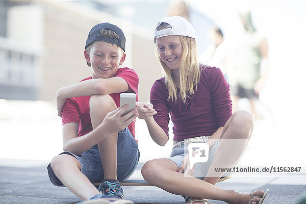 Junge und Mädchen  die ein Smartphone benutzen  sitzen auf dem Skateboard.