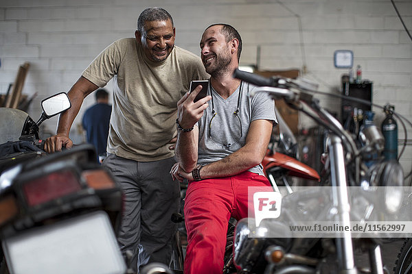 Zwei lächelnde Mechaniker in der Motorradwerkstatt beim Blick aufs Handy