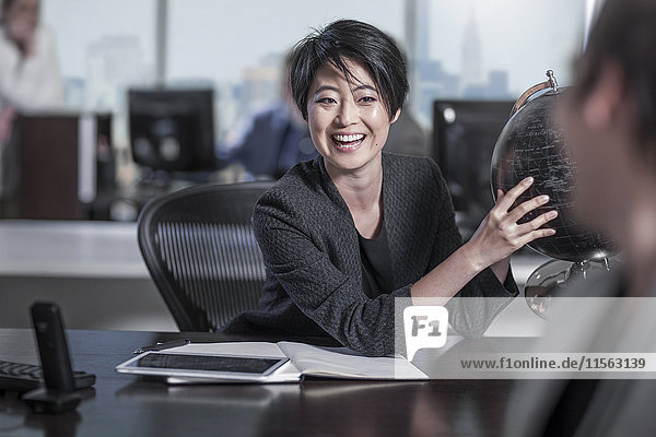 Lachende Frau mit Globus sitzt am Schreibtisch im Stadtbüro
