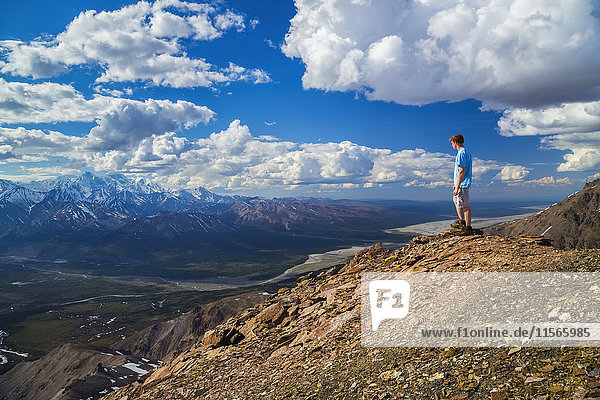 'A hiker stands on a rocky ledge overlooking the Delta River valley in an area of the Alaska Range known as the Delta Mountains; Alaska  United States of America'