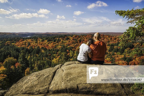 'Couple sitting on a cliff overlooking the fall colours in Algonquin Park  woman has head on man's shoulder; Ontario  Canada'