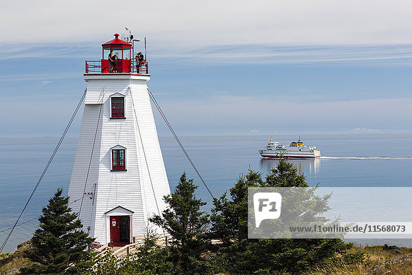 Weißer Leuchtturm mit Menschen auf der obersten Plattform mit Blick auf die Fährüberfahrt in der Ferne und den dunstigen blauen Himmel; North Head  New Brunswick  Kanada'.