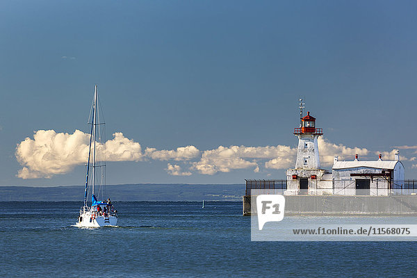 Weißes Segelboot im Wasser neben einem weiß-roten Leuchtturm am Ende eines Piers mit blauem Himmel und Wolken in der Ferne; Port Colborne  Ontario  Kanada'.