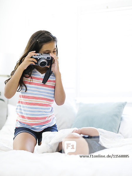 Girl (10-11) taking photo of her small brother (12-17 months) lying on bed