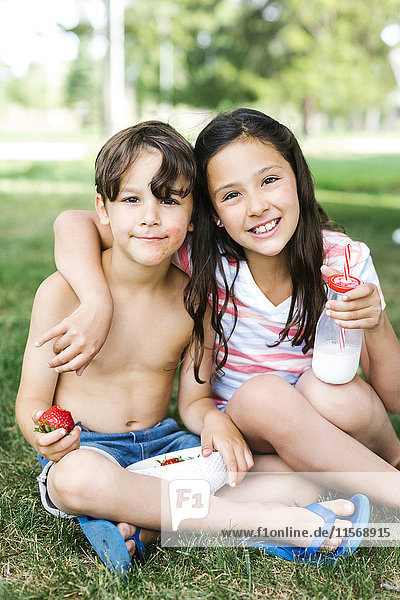 Boy (6-7) and girl (10-11) sitting arm around on grass in summer