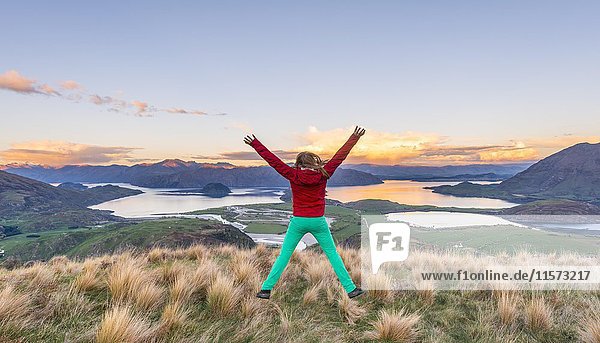 Wanderer springt mit ausgestreckten Gliedmaßen in die Luft  Blick auf Lake Wanaka und Berge  Sonnenuntergang  Rocky Peak  Glendhu Bay  Otago  Southland  Neuseeland  Ozeanien