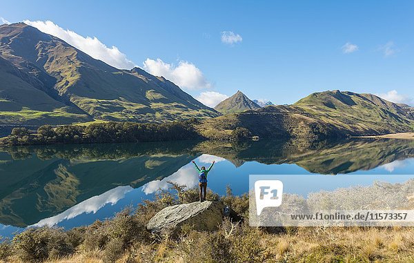 Wanderer steht auf Felsen und streckt die Arme aus  Berge spiegeln sich im See  Moke Lake in der Nähe von Queenstown  Region Otago  Southland  Neuseeland  Ozeanien