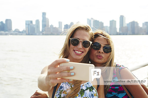 Two women taking smartphone selfie on waterfront with skyline  New York  USA