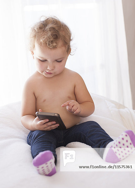 Weibliches Kleinkind sitzt auf dem Bett und nutzt den Touchscreen eines Smartphones