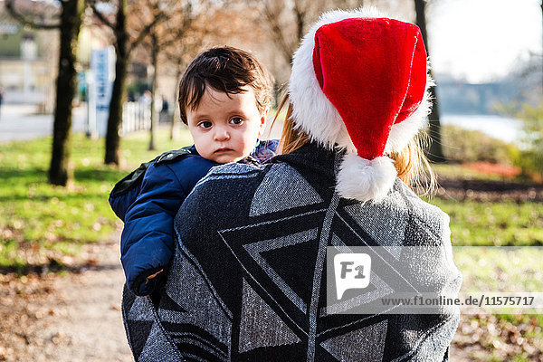 Über-Schulter-Ansicht einer jungen Frau mit Weihnachtsmannhut  die einen kleinen Jungen im Park trägt