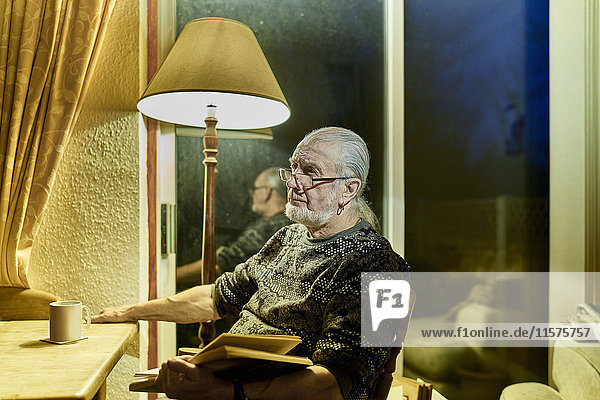 Älterer Mann mit Buch träumt in der Abenddämmerung im Wohnzimmer