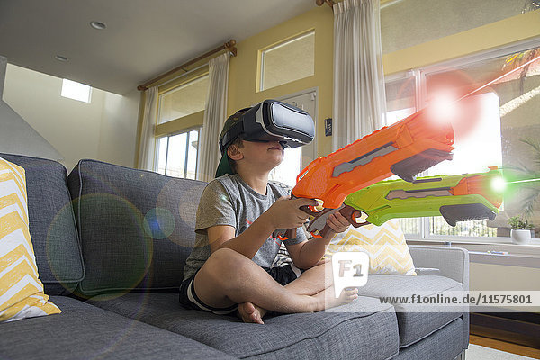 Junge Junge sitzt im Schneidersitz auf dem Sofa  trägt ein Virtual-Reality-Headset  hält Laserpistolen  digitales Komposit
