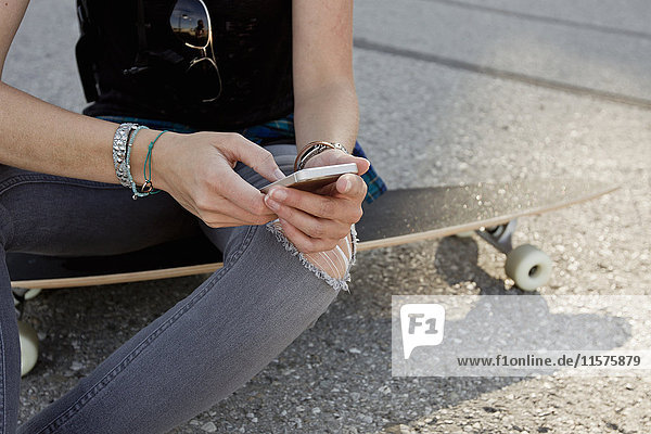 Mid section of female skateboarder sitting on skateboard texting on smartphone