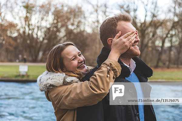 Junge Frau bedeckt die Augen ihres Freundes am Flussufer des Parks