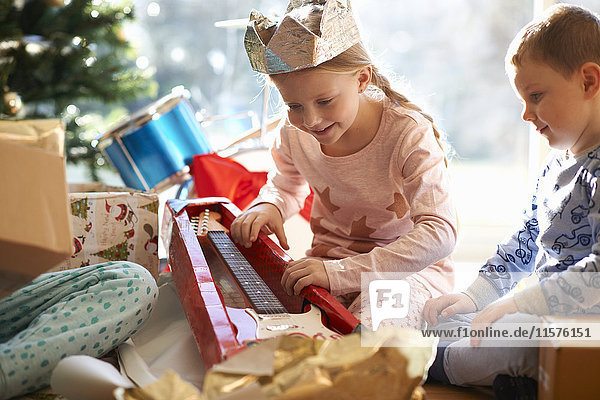 Mädchen und Bruder auf dem Wohnzimmerboden mit Blick auf ein Weihnachtsgeschenk für eine Spielzeuggitarre