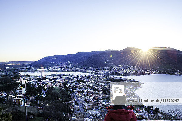 Rückansicht eines Jungen  der den Sonnenuntergang über den Bergen und der Stadt am See beobachtet  Italien