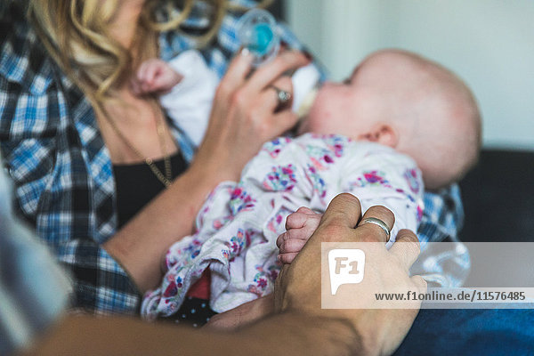 Mother and father sitting on sofa with baby daughter mother feeding baby with bottle mid section
