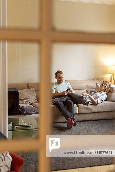 Mid adult couple relaxing on sofa  man using smartphone  woman using digital tablet