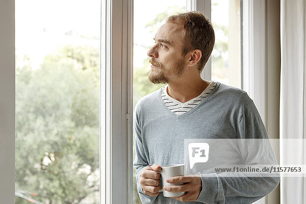 Mann zu Hause  schaut aus dem Fenster  hält heißes Getränk in der Hand