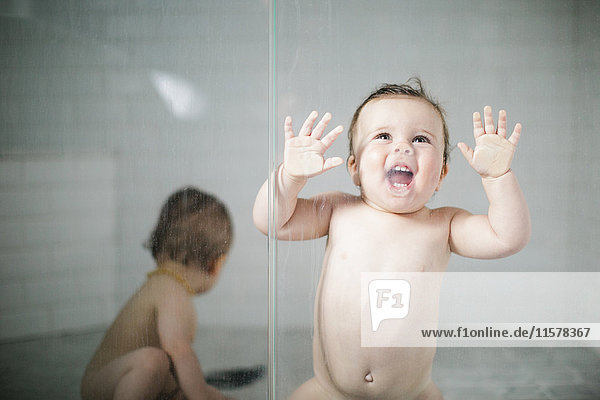 Naked toddler leaning against glass door in shower