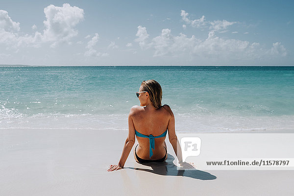 Rückansicht einer Frau im Bikini  die am Strand sitzt und auf das blaue Meer blickt  Anguilla  Saint Martin  Karibik