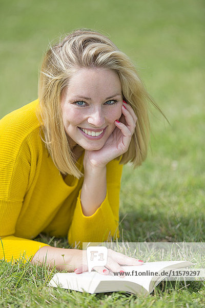 Blonde hübsche Frau beim Lesen eines Buches im Park lächelnd vor der Kamera