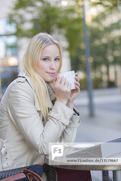 Hübsche blonde Frau genießt eine Tasse Kaffee in der Innenstadt mit Blick auf die Kamera.