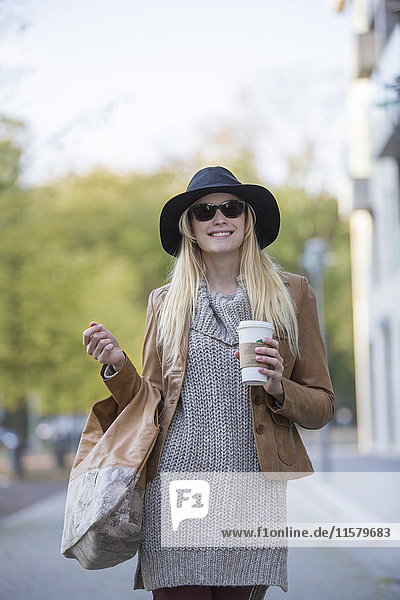 Hübsche blonde Frau mit Hut  Sonnenbrille und Kaffee zum Spazierengehen im Stadtzentrum.