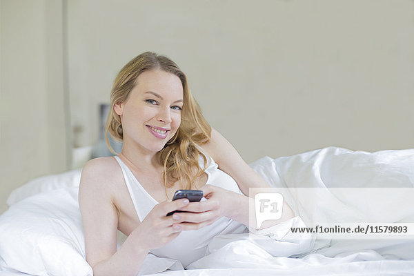 Content pretty blonde woman with mobile in bed smiling at camera