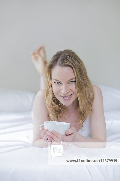 Hübsche blonde Frau mit einer Tasse Tee im Bett  lächelnd vor der Kamera.
