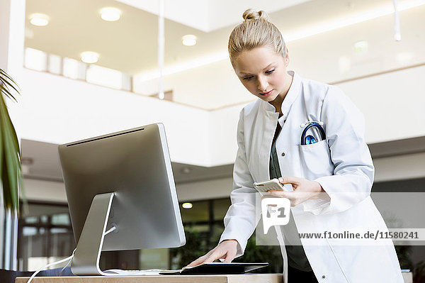 Doctor standing beside computer  holding open diary  looking at smartphone