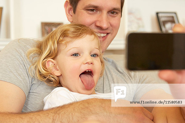 Female toddler sitting on father's knee sticking out tongue for smartphone selfie