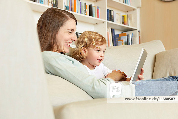 Female toddler sitting on sofa with mother looking at digital tablet
