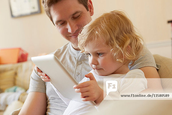 Weibliches Kleinkind sitzt mit seinem Vater auf dem Sofa und benutzt ein digitales Tablett