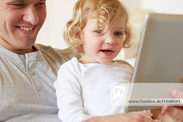 Female toddler sitting on father's knee using digital tablet