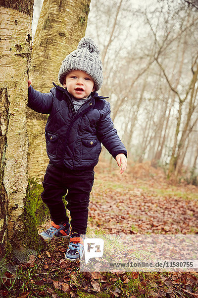 Portrait of male toddler in knitted hat leaning against forest tree