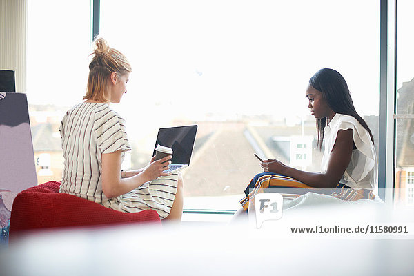 Zwei Businessfrauen sitzen auf Sitzsäcken und schauen auf Laptop und Smartphone