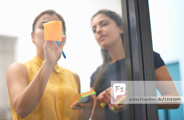 Zwei Geschäftsfrauen kleben Klebezettel an Büro-Glaswand