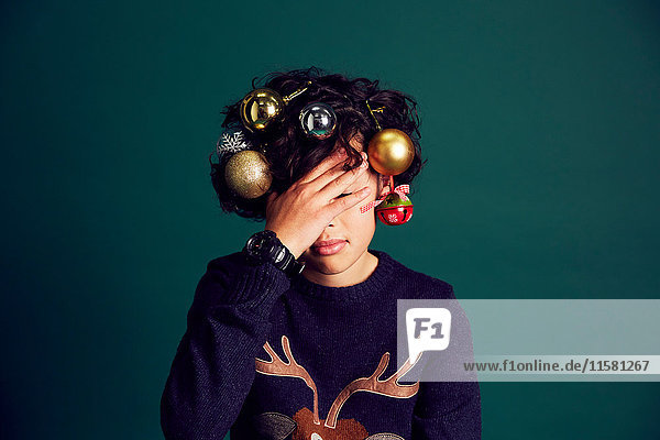 Teenager-Junge mit Weihnachtspullover und Christbaumkugeln im Haar  das Gesicht mit der Hand bedeckt