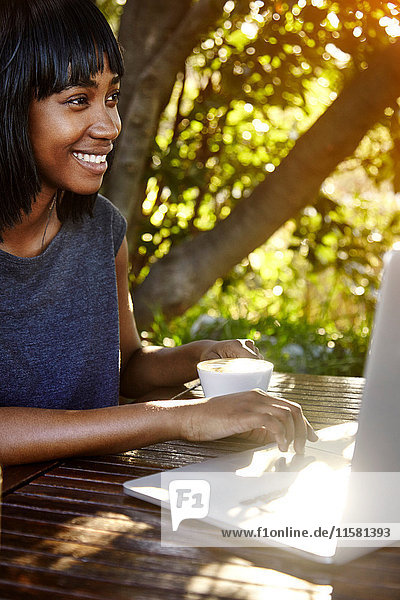 Junge Frau sitzt im Freien  benutzt Laptop  trinkt Kaffee