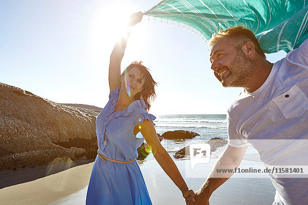 Erwachsenes Paar hält am Strand Händchen  Frau hält Schal in der Luft