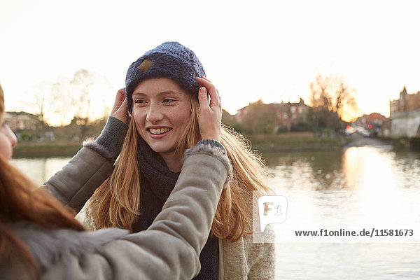 Young woman putting knitted hat on friend  outdoors  smiling
