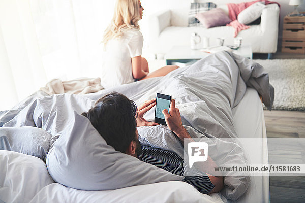 Man in bed using smartphone  woman sitting at end of bed
