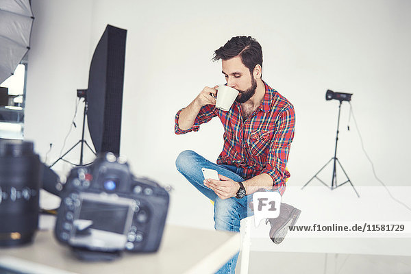 Male photographer drinking coffee and looking at smartphone in studio