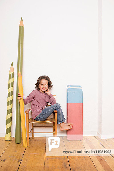 Junges Mädchen sitzt auf einem Stuhl,  hält einen Bleistift in Riesengröße in der Hand,  neben sich an der Wand lehnt ein riesengroßes Briefpapier