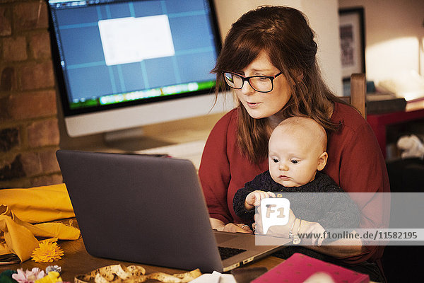 Eine Frau sitzt mit einem Baby auf dem Schoß  das einen Computer-Laptop benutzt  wobei beide Personen aufmerksam auf den Bildschirm schauen.