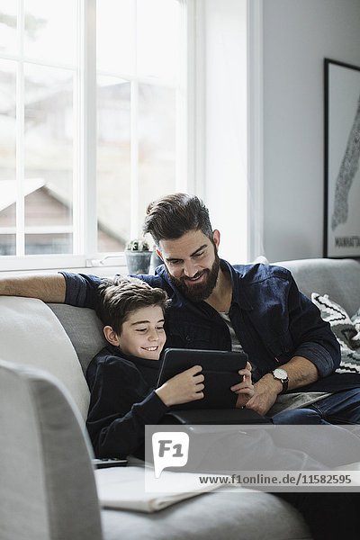 Glücklicher Vater sieht den Jungen mit dem digitalen Tablett an  während er im Wohnzimmer auf dem Sofa sitzt.