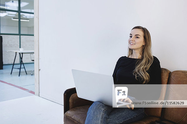 Lächelnde junge Frau schaut weg  während sie mit dem Laptop auf dem Sofa im Büro sitzt.
