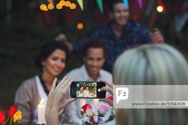Abgeschnittenes Bild einer Frau  die glückliche Freunde durch ein Smartphone auf einer Gartenparty fotografiert.